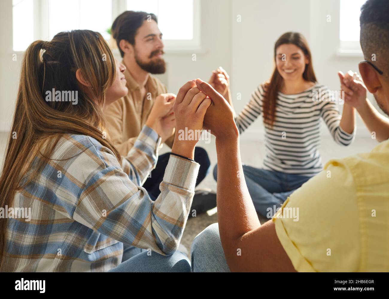 Gruppo multietnico di amici felici seduti in cerchio sul pavimento, tenendo le mani e sorridendo Foto Stock