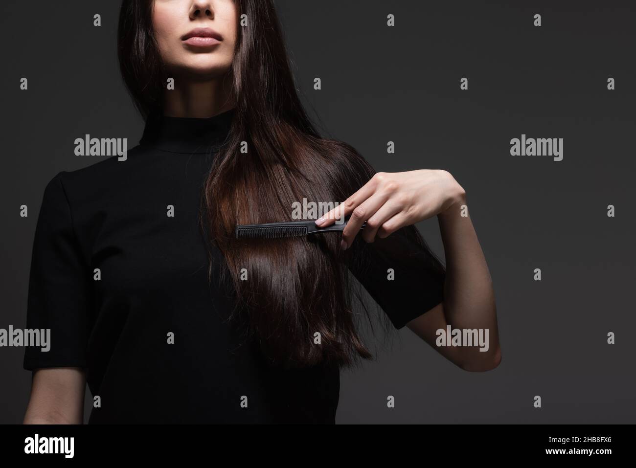 vista ritagliata della giovane donna spazzolando lunghi capelli luminosi con pettine isolato su grigio scuro Foto Stock