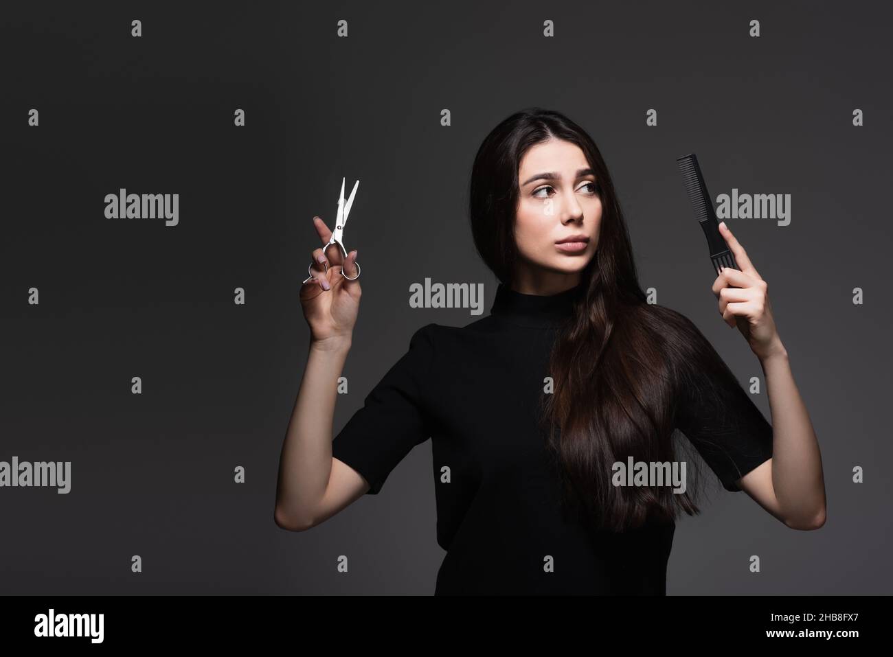 giovane donna con capelli lunghi e lucenti che tengono forbici e pettine isolato su grigio scuro Foto Stock