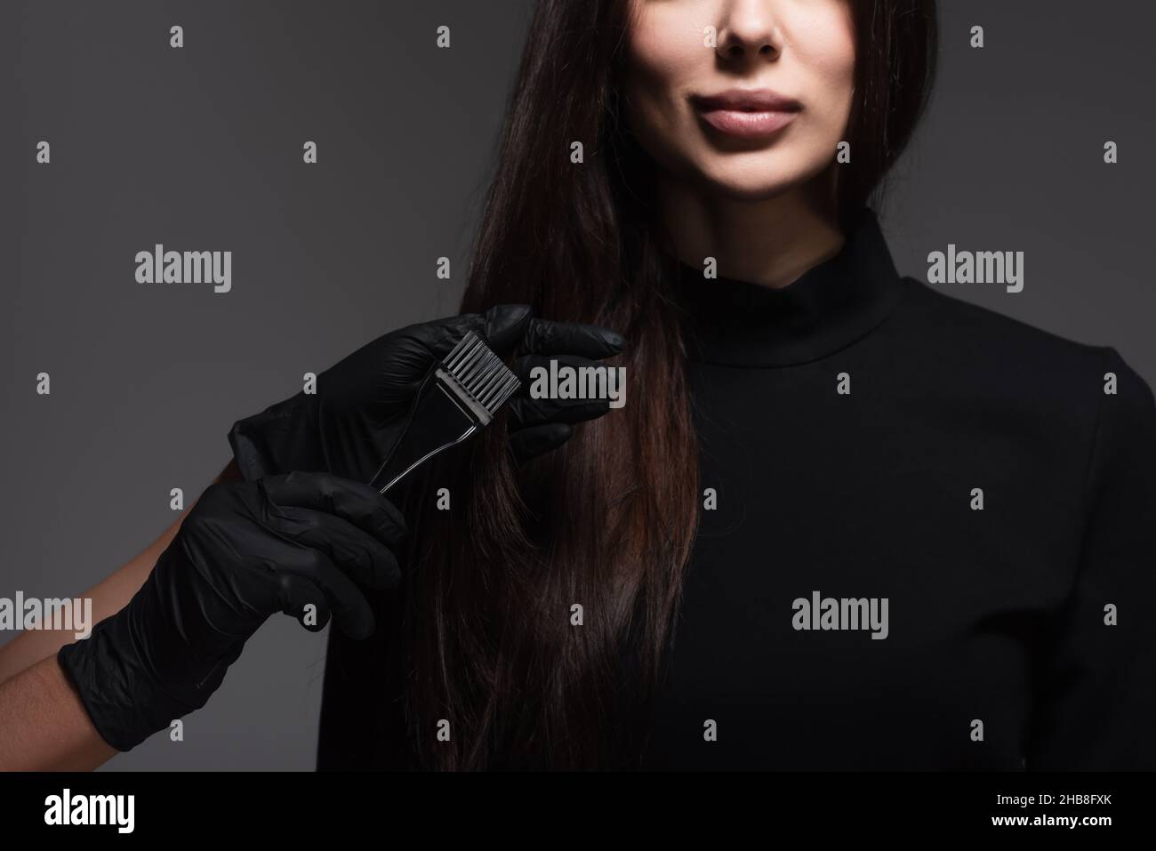 vista croppata di spazzola di tenuta di hairstylist vicino alla donna con capelli marroni isolati su grigio scuro Foto Stock