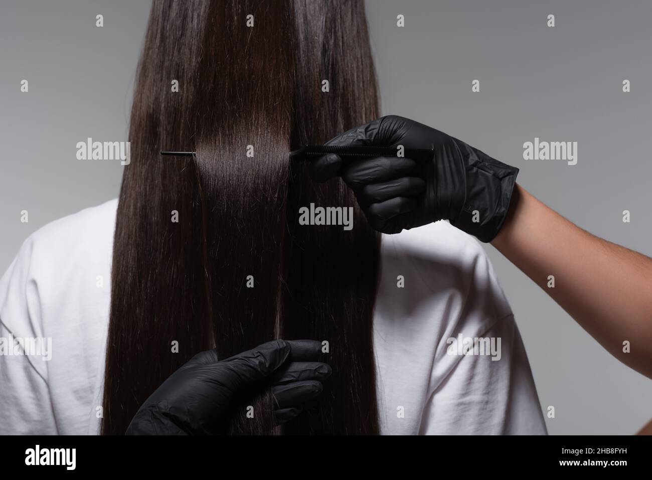 vista posteriore della giovane donna con capelli lisci lunghi vicino al colorist in lattice guanti isolati sul grigio Foto Stock