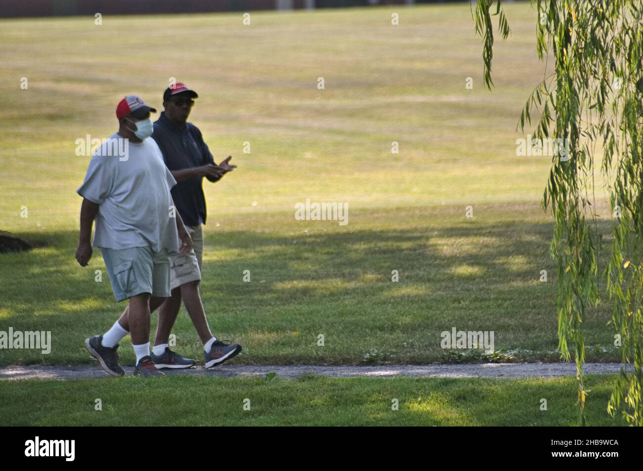 Due uomini neri afroamericani di mezza età, uno con maschera e uno senza maschera, camminando in un parco locale Foto Stock