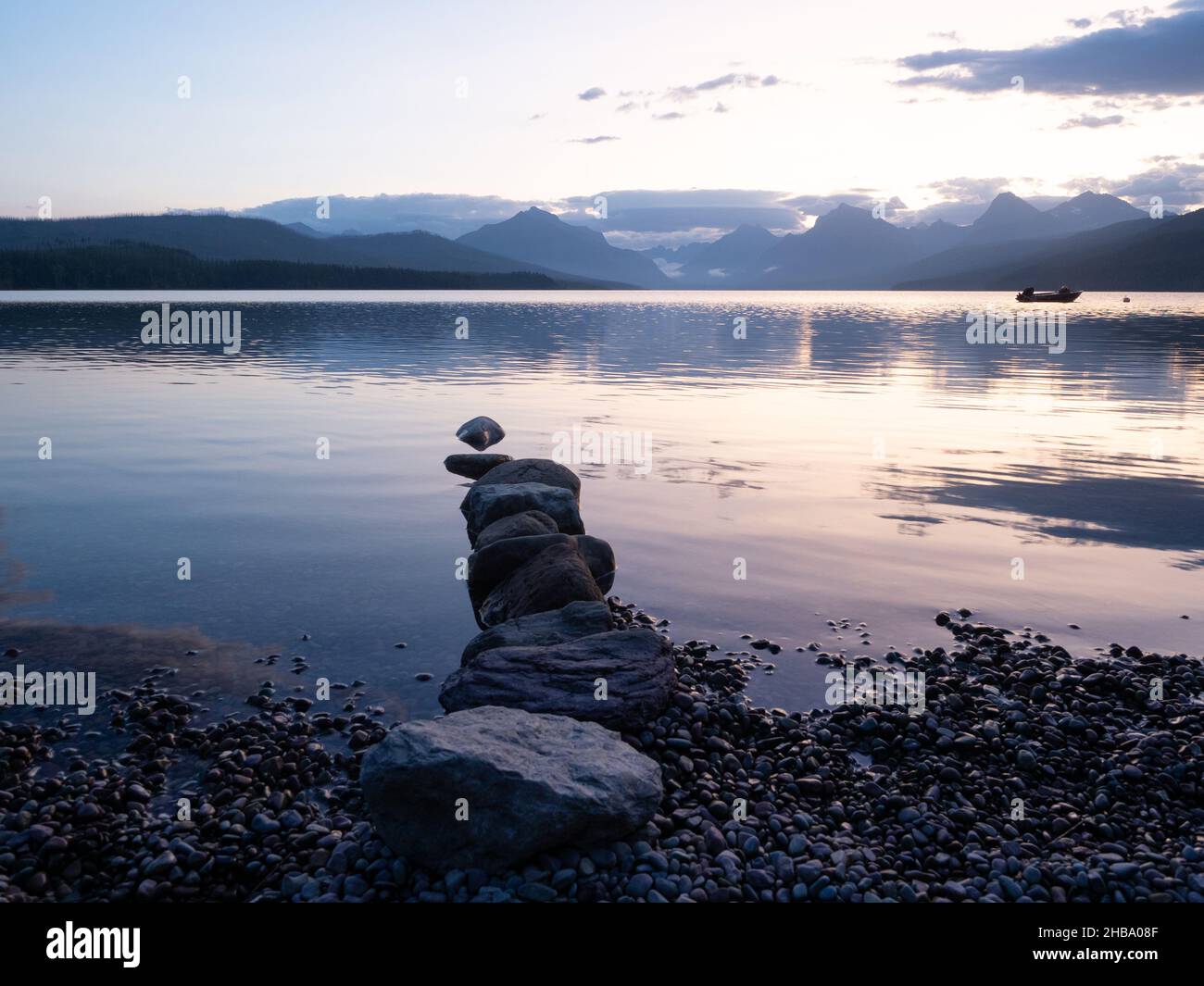 Una linea di pietre che si aggettano nel Lago McDonald nel Parco Nazionale del Ghiacciaio con l'aspra catena montuosa Livingston e una piccola barca a motore nel backgroun Foto Stock