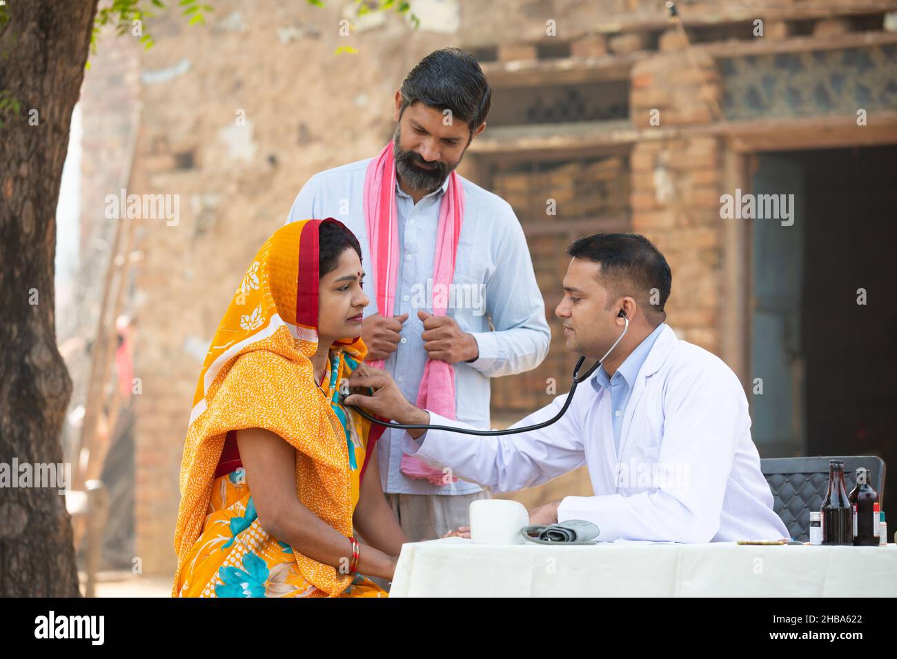 Medico indiano maschio con stetoscopio di controllo del battito cardiaco paziente o respiro al villaggio, donna che indossa sari con suo marito che ottiene esame da medico p Foto Stock