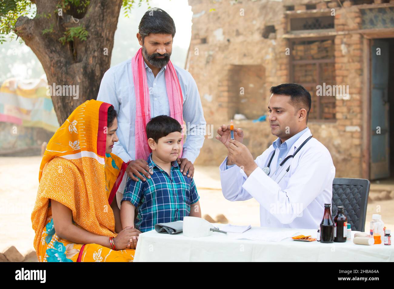 Medico indiano con siringa o iniezione medica in mano, bambino piccolo paziente in villaggio, donna che indossa sari con suo marito e figlio seduto accanto t. Foto Stock