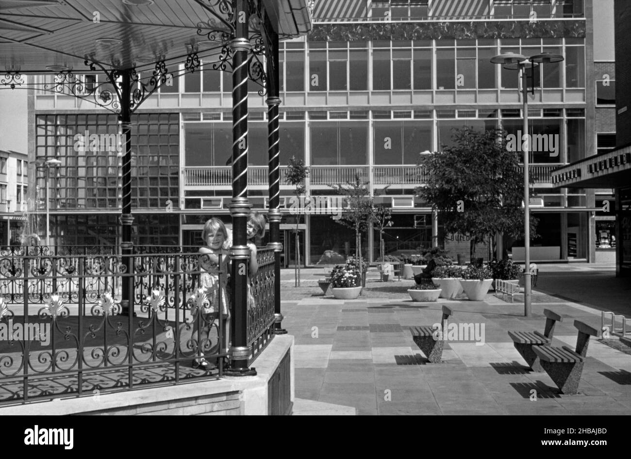 Queen’s Square, Crawley ‘New Town’, West Sussex, Inghilterra, Regno Unito con due ragazze che giocano sul suo iconico banco da banda fotografato nel 1960s. L’espansione delle principali strutture commerciali era essenziale per l’espansione di Crawley. La prima tappa fu la Broadwalk nel 1954, dopo l'apertura della Piazza della Regina da parte di sua Maestà la Regina nel 1958. Dopo la seconda guerra mondiale, al fine di delocalizzare coloro che vivono nelle abitazioni povere o bombardate di Londra, un gran numero di persone e di posti di lavoro sono stati spostati in nuove città intorno alla se Inghilterra e Crawley è stata la prima di queste – una fotografia d’epoca del 1960s. Foto Stock