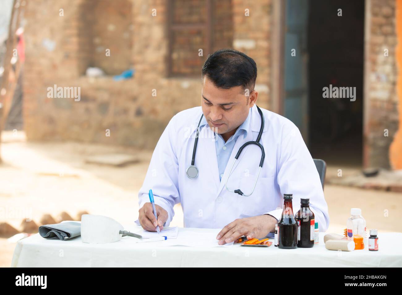 Giovane medico indiano maschio che scrive la prescrizione su carta mentre si siede all'ospedale esterno del villaggio della scrivania. Assistenza sanitaria rurale. Foto Stock