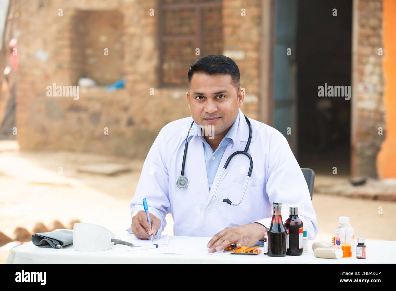 Ritratto di giovane medico indiano maschio che scrive la prescrizione su carta mentre si siede all'ospedale esterno del villaggio della scrivania, guardando la macchina fotografica. Assistenza sanitaria rurale Foto Stock