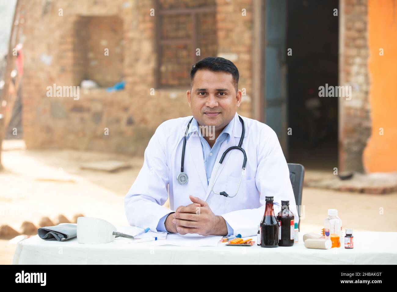 Ritratto di giovane medico indiano maschio seduto alla scrivania ospedale villaggio all'aperto con un sacco di medicina intorno, guardando la macchina fotografica. Assistenza sanitaria rurale Foto Stock