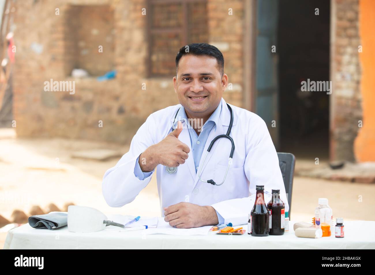 Ritratto di positivo giovane medico indiano maschio fare i pollici in su mentre si siede all'ospedale esterno del villaggio della scrivania con i lotti della medicina intorno, guardando Foto Stock