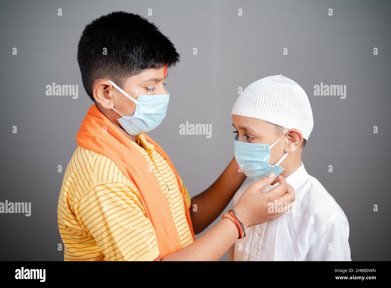 Hindu Kid aiutare il suo amico musulmano a indossare correttamente maschera medica viso su sfondo grigio per proteggere dal coronavirus o covid-19 pandemia - concetto Foto Stock