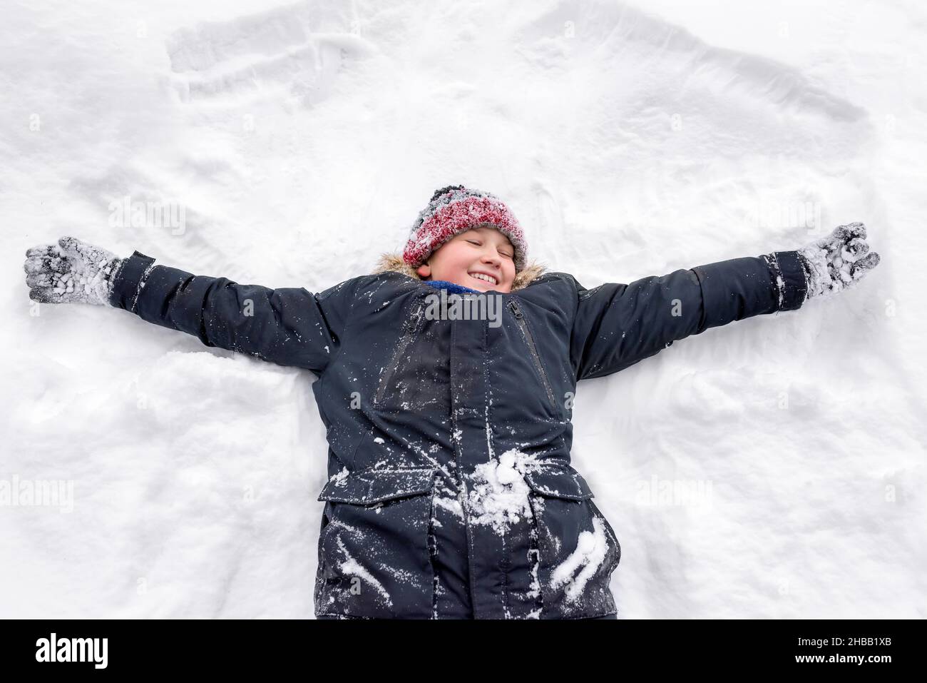 Un ragazzo sorridente in abiti caldi si trova sulla schiena e mostra un angelo nella neve. Foto Stock