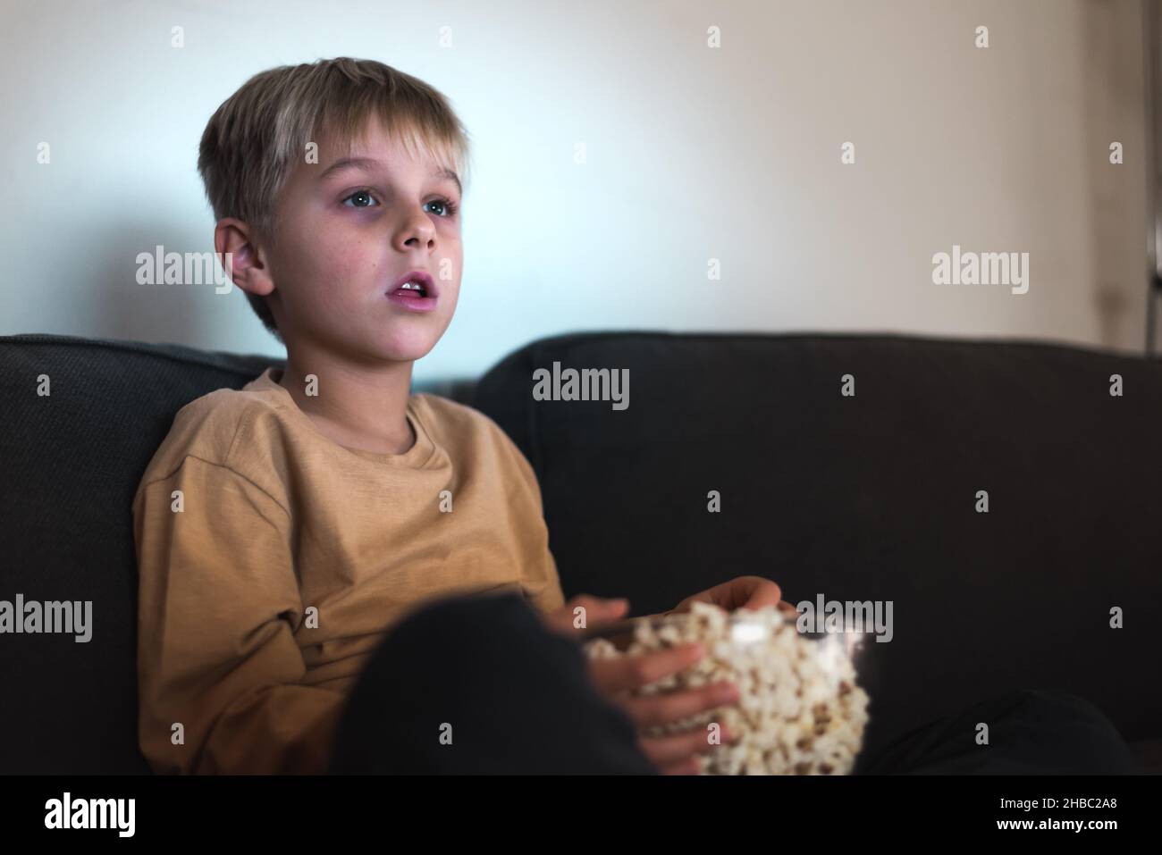 Ragazzo adorabile attento con capelli biondi in abiti casual guardando un interessante film su T. con bocca aperta e mangiare ciotola di popcorn durante il fine settimana a casa Foto Stock