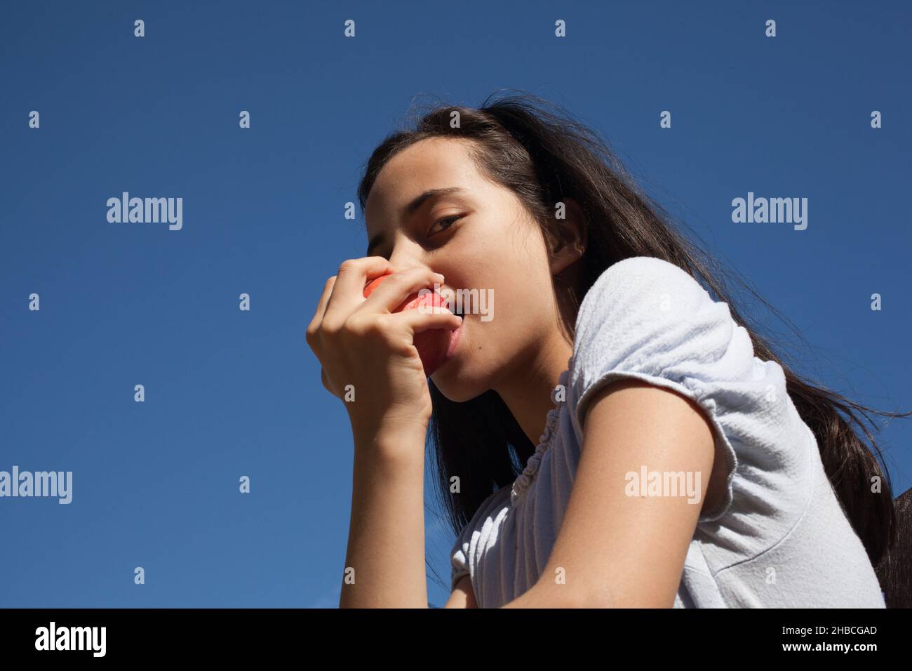 Una bambina di undici anni che mangia una mela rossa con un cielo blu brillante dietro Foto Stock