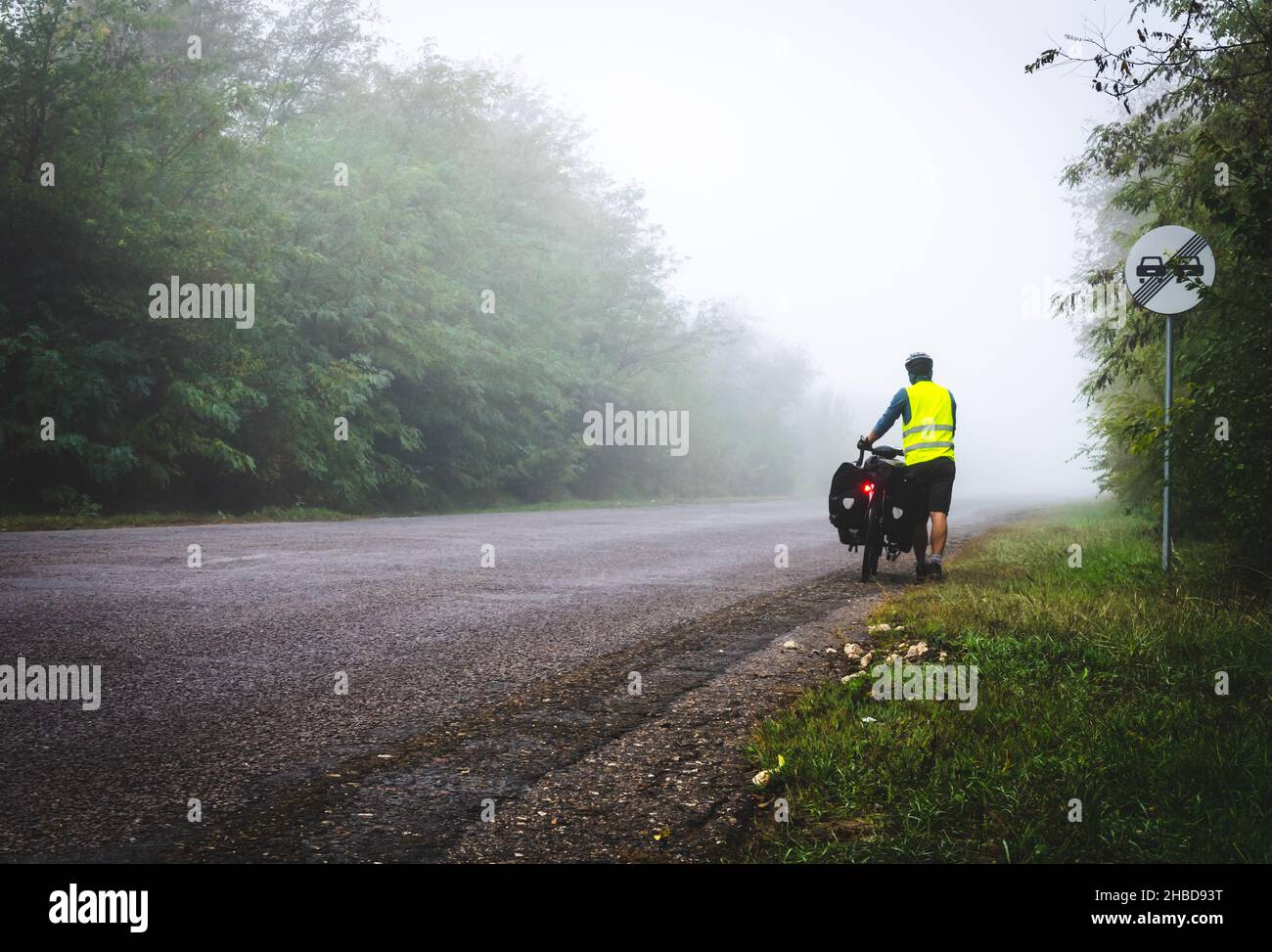 Il ciclista con giubbotto riflettente e accendini si sta camminando lungo la strada per la nebbia in natura. Escursioni in bicicletta in cattive condizioni meteorologiche e cattiva visibilità Foto Stock