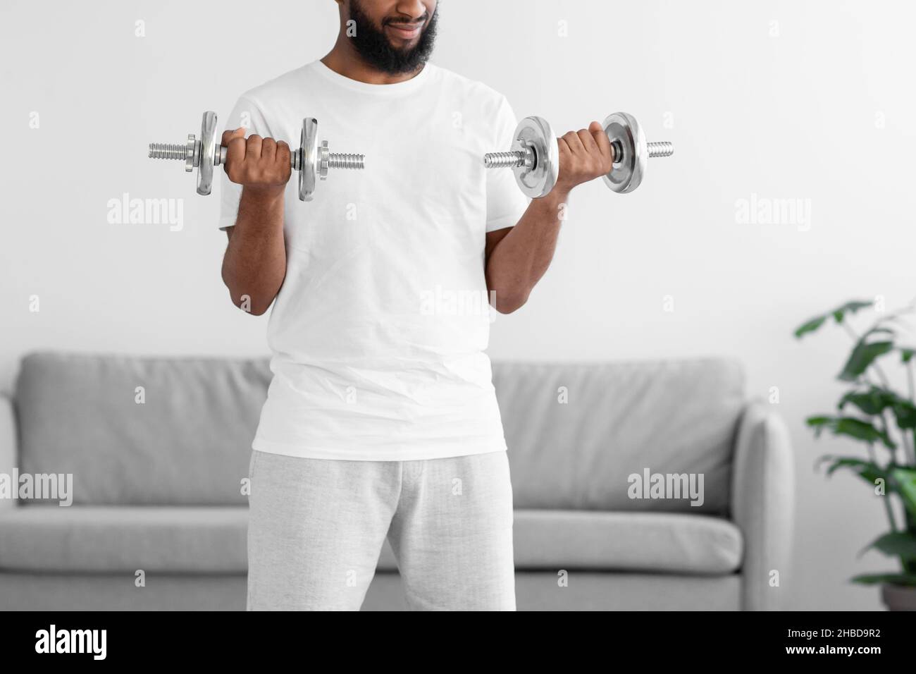 L'atleta nero irriconoscibile in abiti sportivi bianchi si esercita con i manubri in salotto Foto Stock
