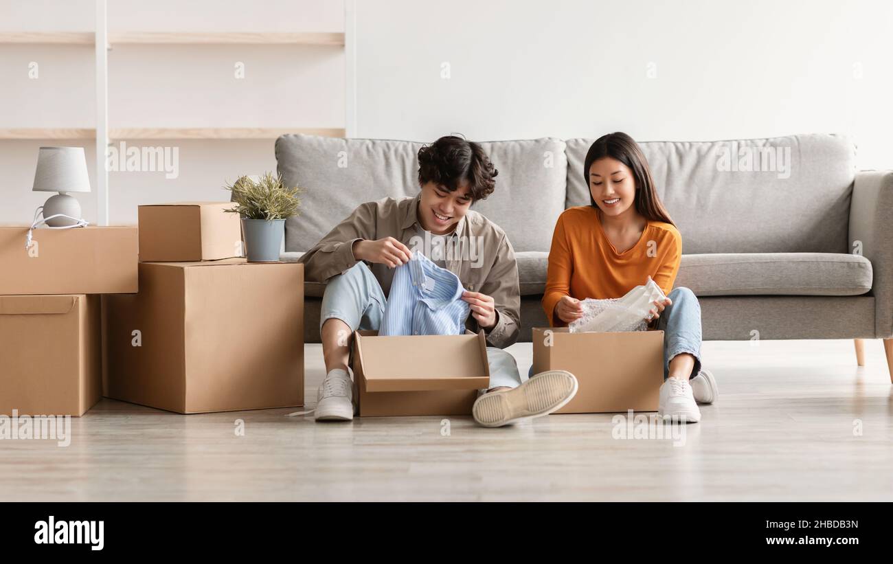 Coppia asiatica gioiosa seduta sul pavimento della nuova casa, disimballaggio delle scatole, spostamento alla loro proprietà, muovendosi insieme Foto Stock