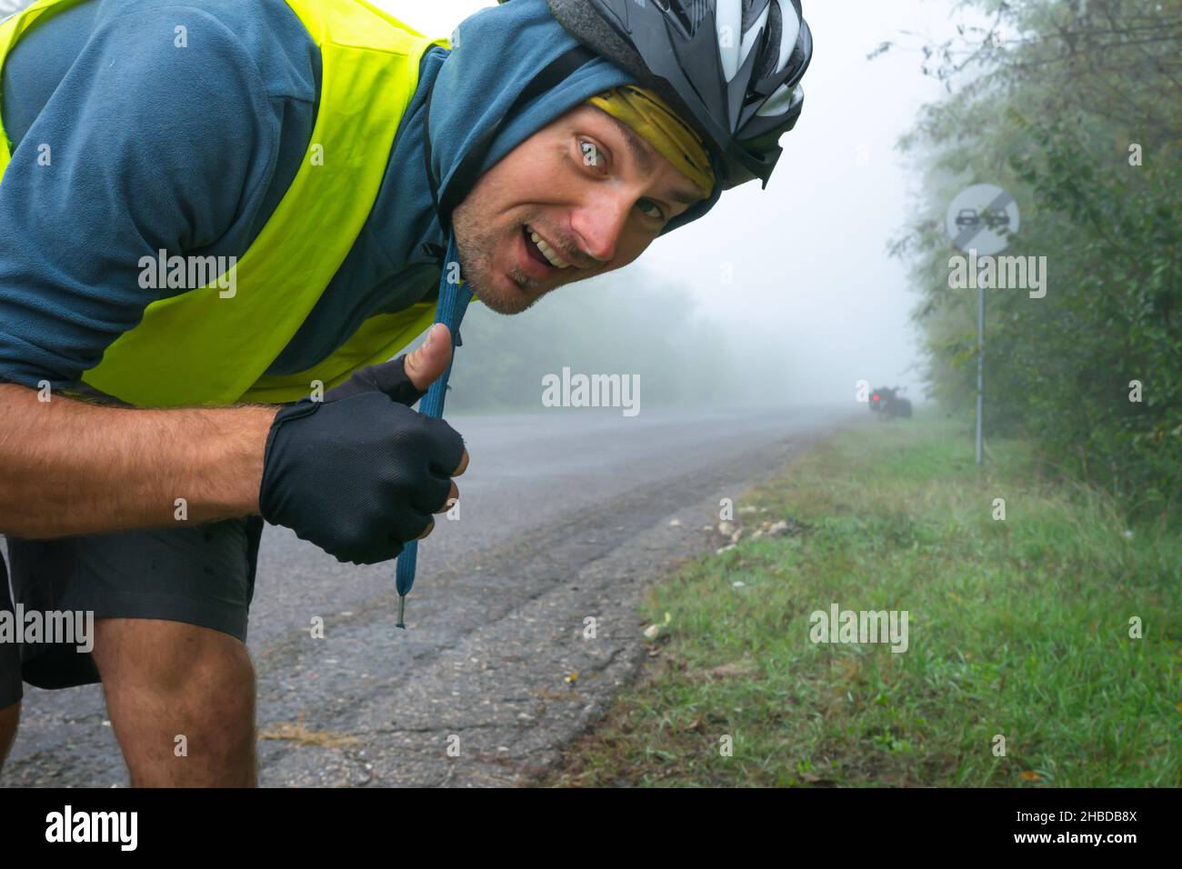 Il ciclista felice con un giubbotto riflettente mostra i pollici in su dalla strada in condizioni di nebbia con un bicyle da turismo nelle vicinanze. Concepton di ciclismo sicuro sotto b Foto Stock