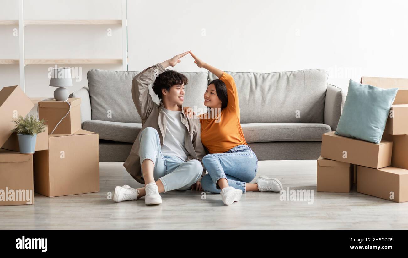 Positivo Asian coppia trasferirsi a casa propria, facendo il movimento casa tetto insieme, seduta sul pavimento tra scatole di cartone Foto Stock