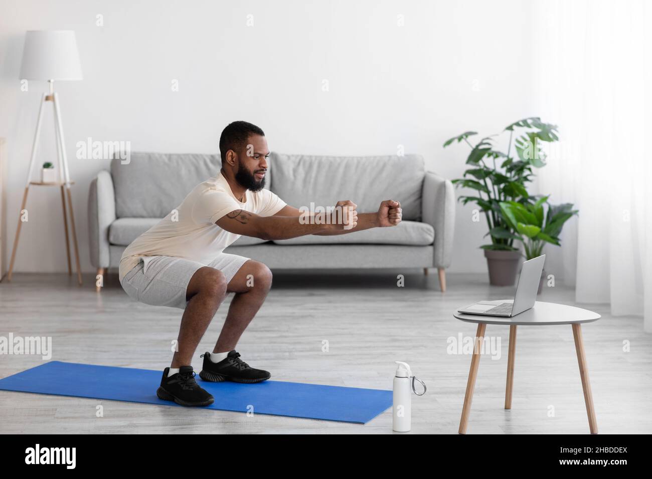 Concentrato giovane atleta nero portico uomo in abiti sportivi bianchi che si accovacciano in salotto interno, profilo Foto Stock