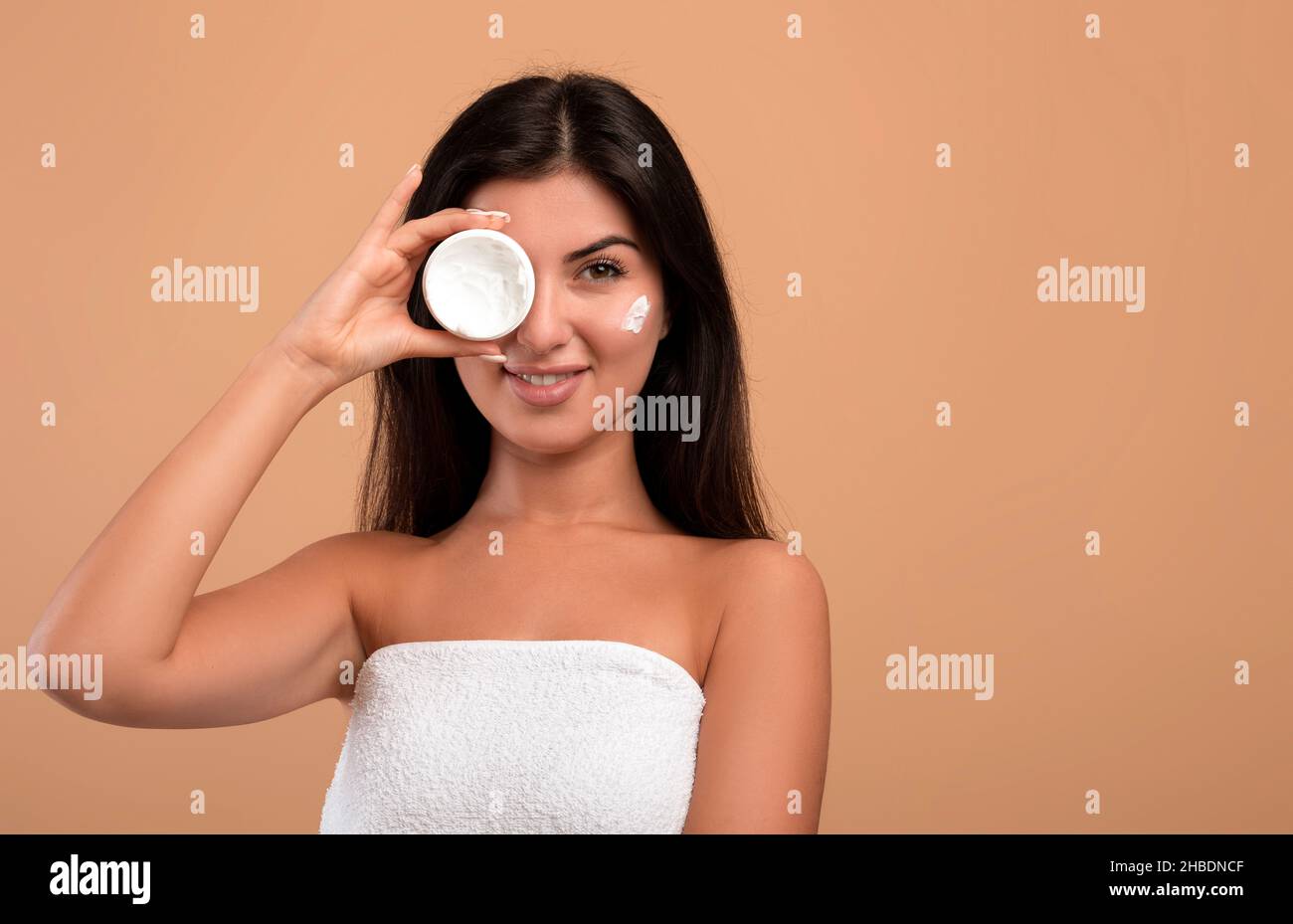 Ritratto di bellezza della graziosa signora armena che tiene il vaso bianco con crema idratante in mano, coprendo un occhio Foto Stock