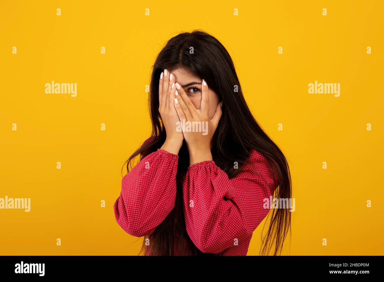 Spaventato giovane armena che cerca di nascondersi, coprendo il viso con le mani e sbirciando con un occhio, sfondo giallo Foto Stock