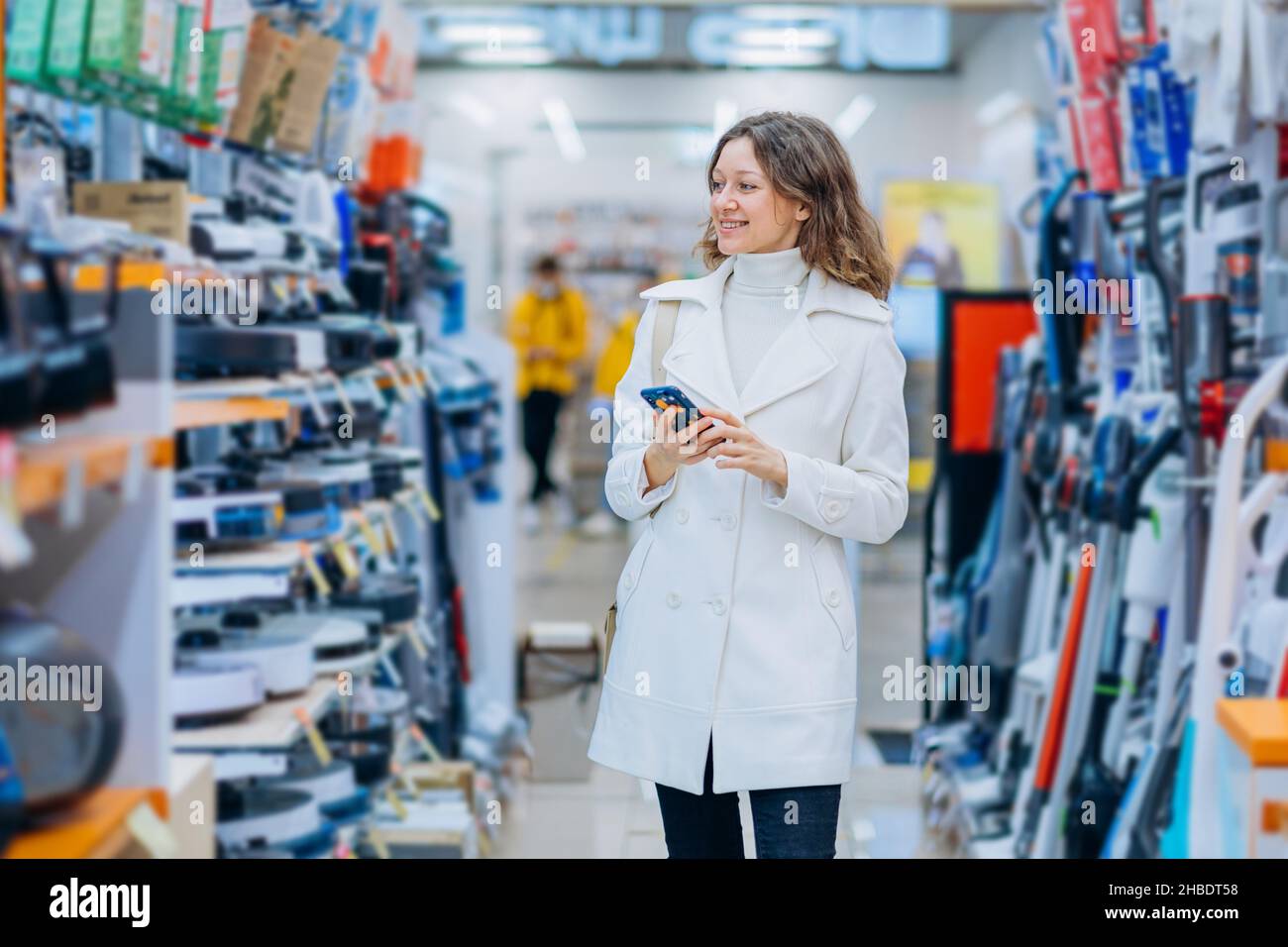 La giovane donna bruna dai capelli ricci indossa un pullover e un cappotto per tenere in mano lo smartphone e sceglie l'attrezzatura per la ristrutturazione con un sorriso luminoso Foto Stock