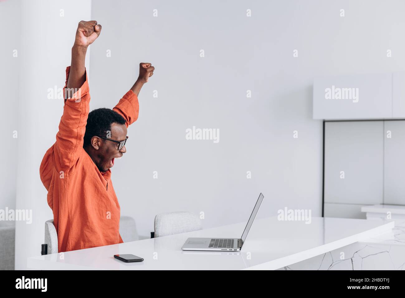 Il giovane uomo afro-americano felice in vestiti colorati e occhiali solleva le mani con gioia guardando il computer portatile al tavolo bianco vista laterale Foto Stock