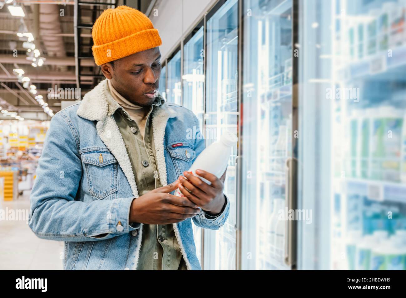 Il ragazzo afro-americano positivo in giacca di denim e cappello arancione prende la bottiglia di latte dalla scatola del frigorifero in vista laterale del moderno supermercato Foto Stock