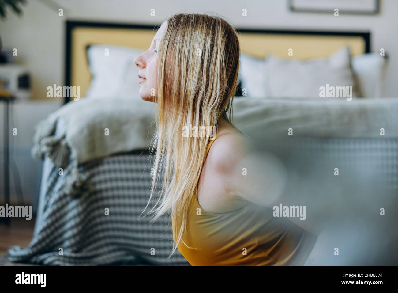 Giovane donna con lunghi capelli biondi in giallo sport top fa esercizi vicino al letto coperto con coperta a righe con cuscini in cima Foto Stock
