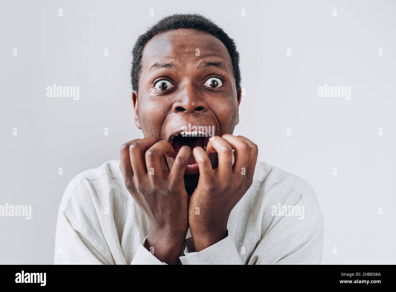 Ritratto di un uomo africano terrorizzato fissando in orrore la macchina fotografica e mettendo le sue mani alla bocca in una camicia di lino chiaro su sfondo bianco. Sorpresa o paura molto forte, orrore negli occhi. Foto Stock