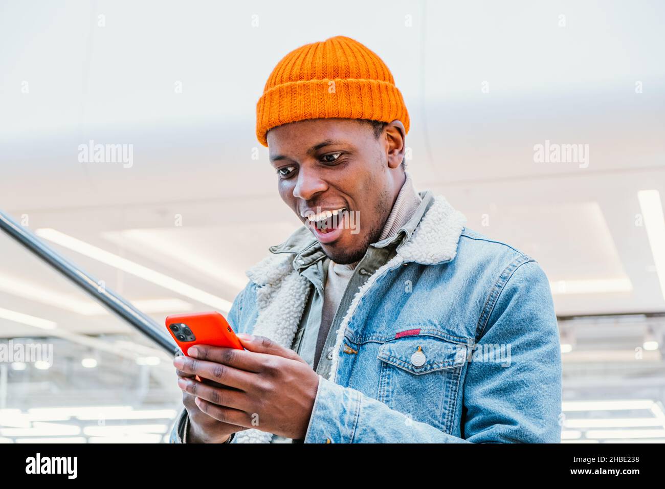 Emozionale felice africano-americano ragazzo in elegante vestito guarda il telefono cellulare che va su scala mobile nel centro commerciale vicino vista Foto Stock