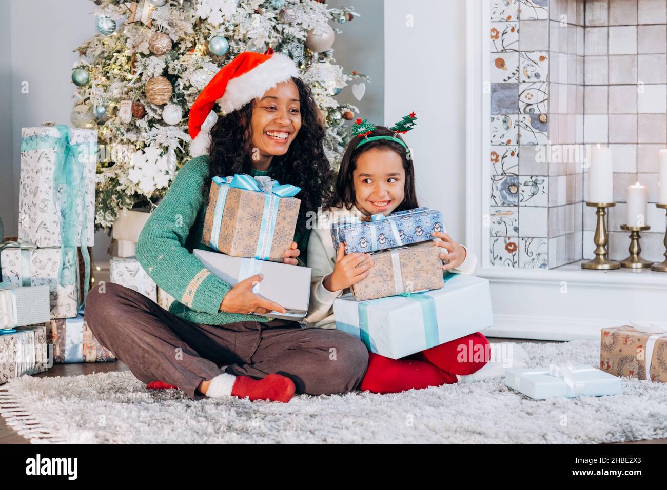 Curly African American donna e bruna ragazza dai capelli lunghi che indossa i cappelli di festa aprire le scatole presenti con sorriso che siede all'albero di Natale Foto Stock