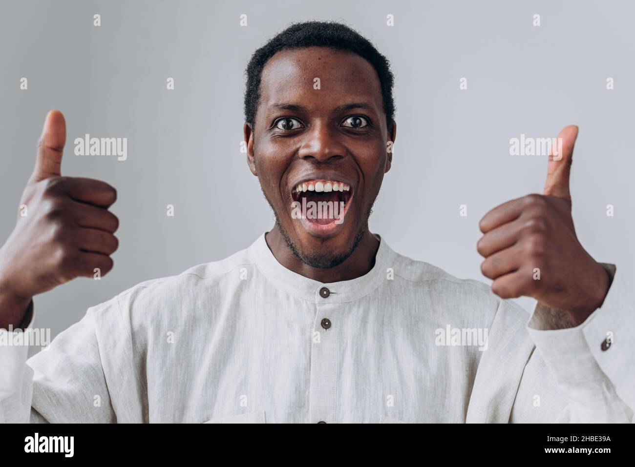 Ritratto di emozionale felice africano-americano ragazzo in camicia bianca mostrando i pollici in posa su sfondo grigio chiaro vista ravvicinata Foto Stock