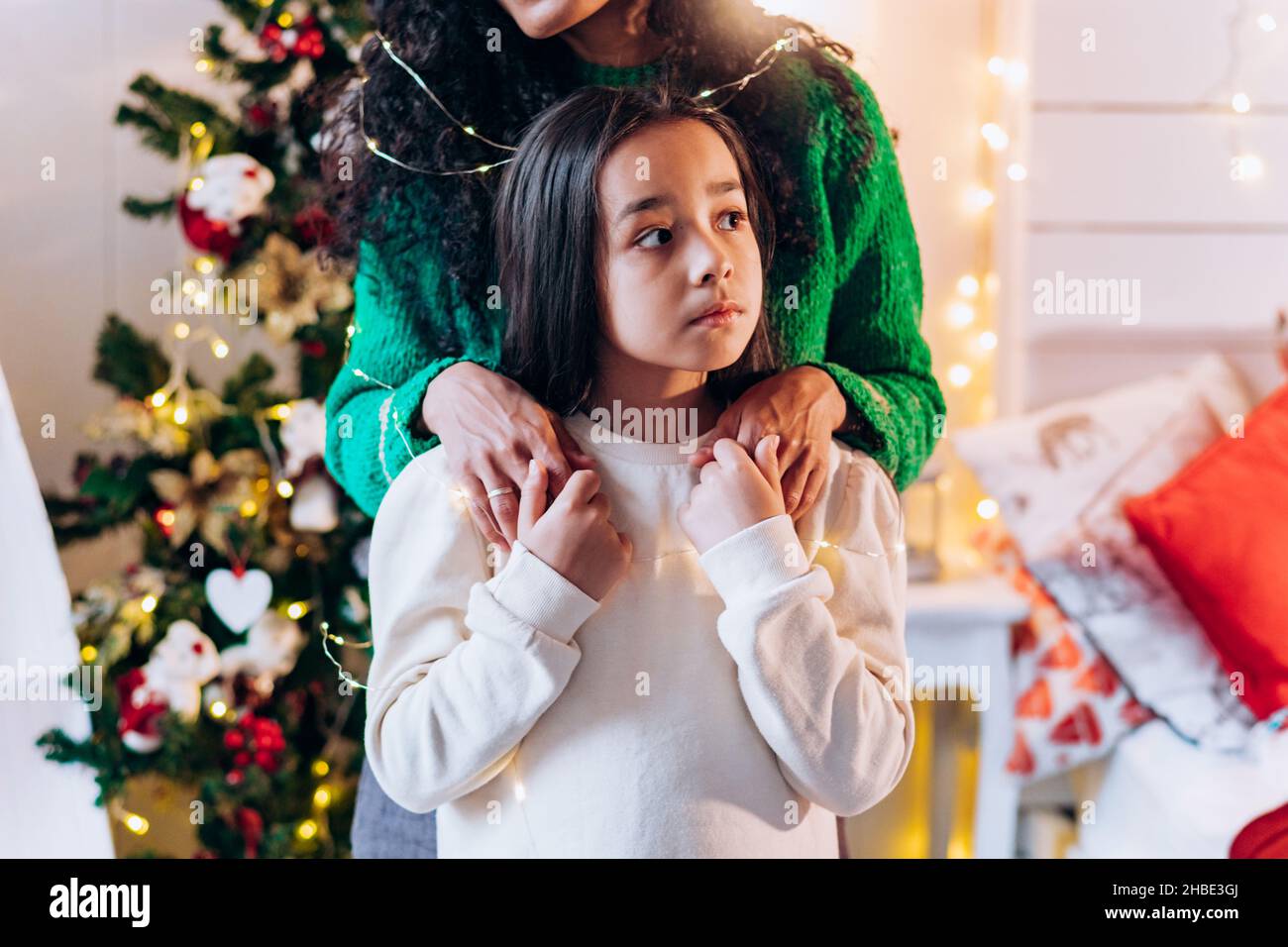 Ritratto di una bambina e sua madre in piedi dietro di lei in un maglione  verde, mettendo le mani sulle sue spalle sullo sfondo di un albero di Natale  con una ghirlanda,