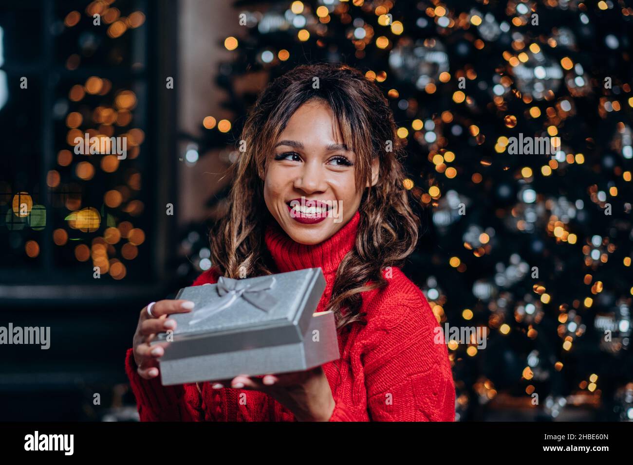 Ritratto di una donna da corsa mista sorridente in un maglione rosso che tiene un regalo aperto sullo sfondo del primo piano albero di natale, guardando da sogno a lato nel primo piano di nuovo anno. Foto Stock