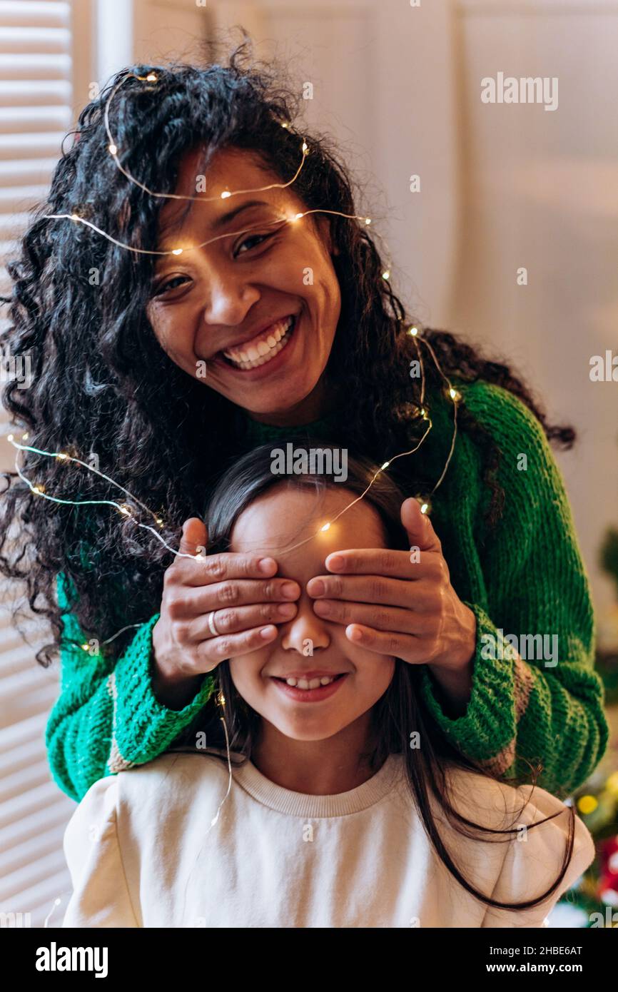 Bruna scolgirl e curly afroamericana madre posare gli occhi chiudendo con le mani in garland illuminante e sorridere in elegante soggiorno Foto Stock