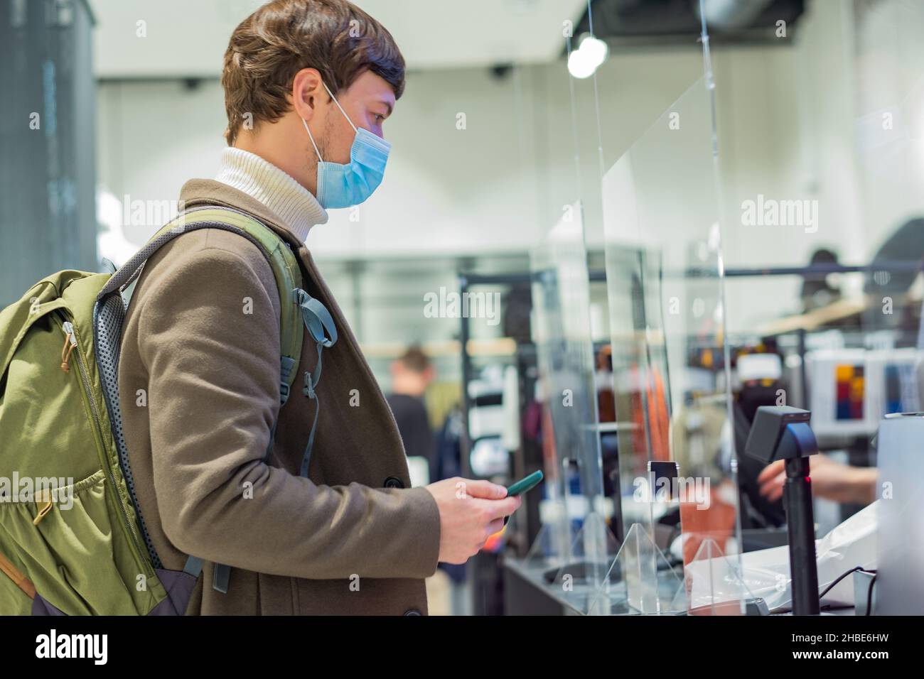 Uomo con capelli corti brunetta in caldo cappotto maglione e maschera medica si alza vicino registratore di cassa tenendo telefono per pagare con pagamento contactless Foto Stock