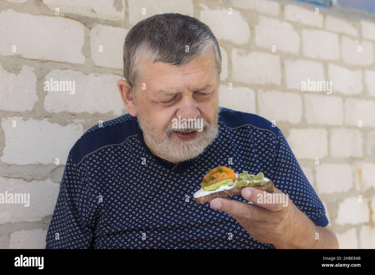 Ritratto di ucraino affamato senior prendere in mano e pronto a mangiare vegan sandwich con cetriolo, pomodoro giallo e guacamole mentre si levano in piedi contro b Foto Stock
