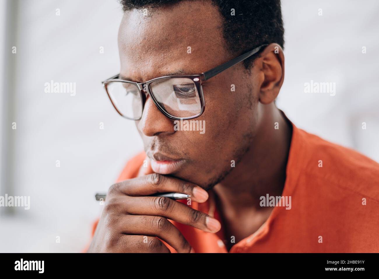 Il giovane uomo afro-americano con occhiali eleganti e penna che indossa abiti arancioni luminosi in camera luminosa vista ravvicinata Foto Stock