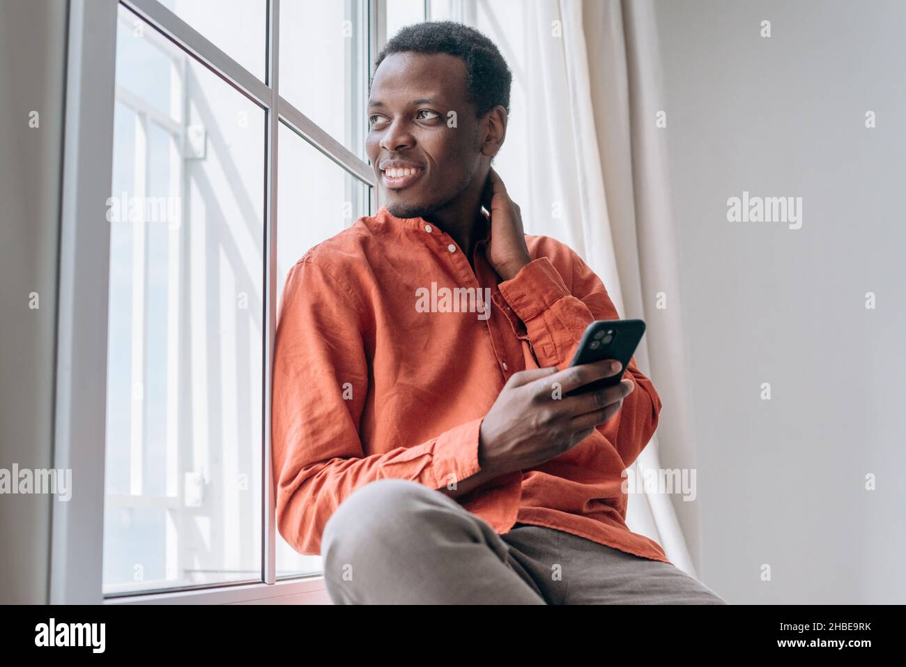 Il ragazzo afro-americano positivo in elegante camicia arancione guarda dalla finestra sorridendo con il telefono cellulare moderno nella stanza luminosa di casa Foto Stock