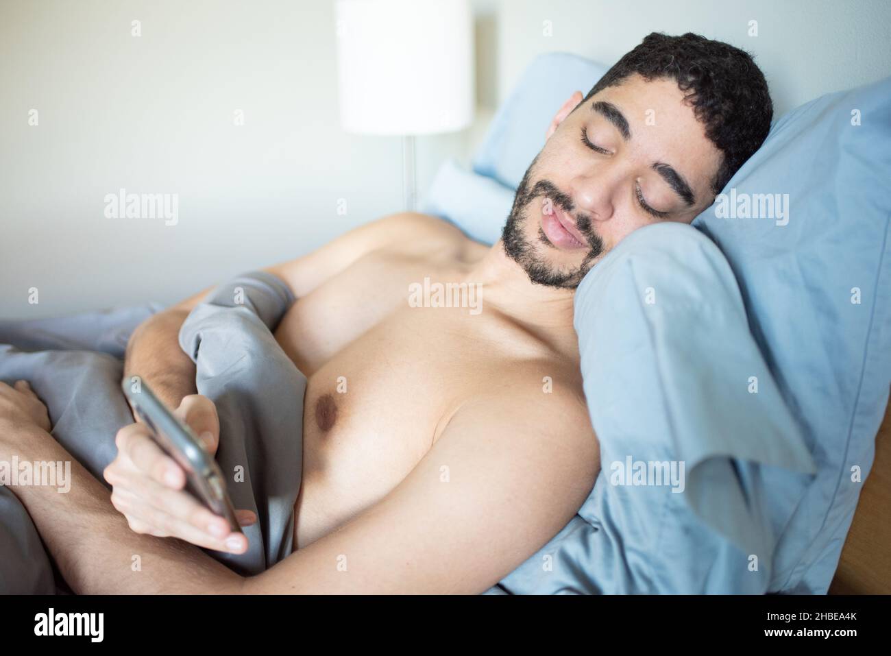 Misto-razza giovane 30-qualcosa uomo millenario steso in letto proppato su cuscini guardando il telefono. Controllare il telefono quando ci si sveglia la mattina Foto Stock