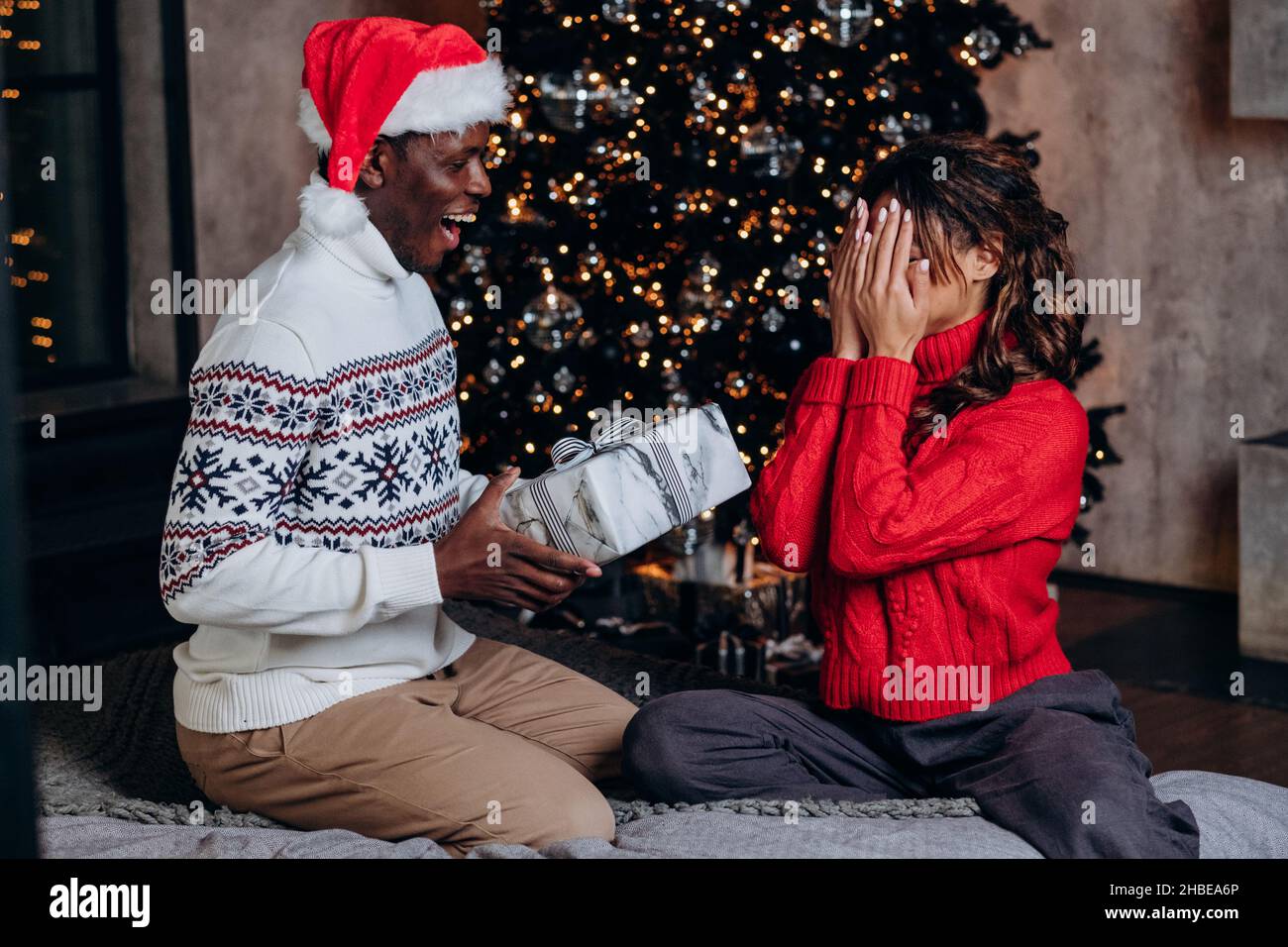 L'uomo afro-americano in un cappello rosso dà un regalo di Natale una scatola sullo sfondo di un albero di Capodanno, una coppia in amore celebra a casa, una donna in un maglione rosso copre il suo volto con le mani ed è in attesa di una sorpresa Foto Stock