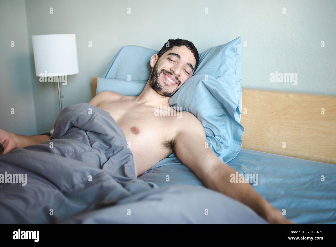 Misto-gara milleniale bearded 30-qualcosa uomo sorride mentre propped in su con due cuscini nel letto con le lenzuola blu chiaro e il comforter grigio, occhi chiusi Foto Stock