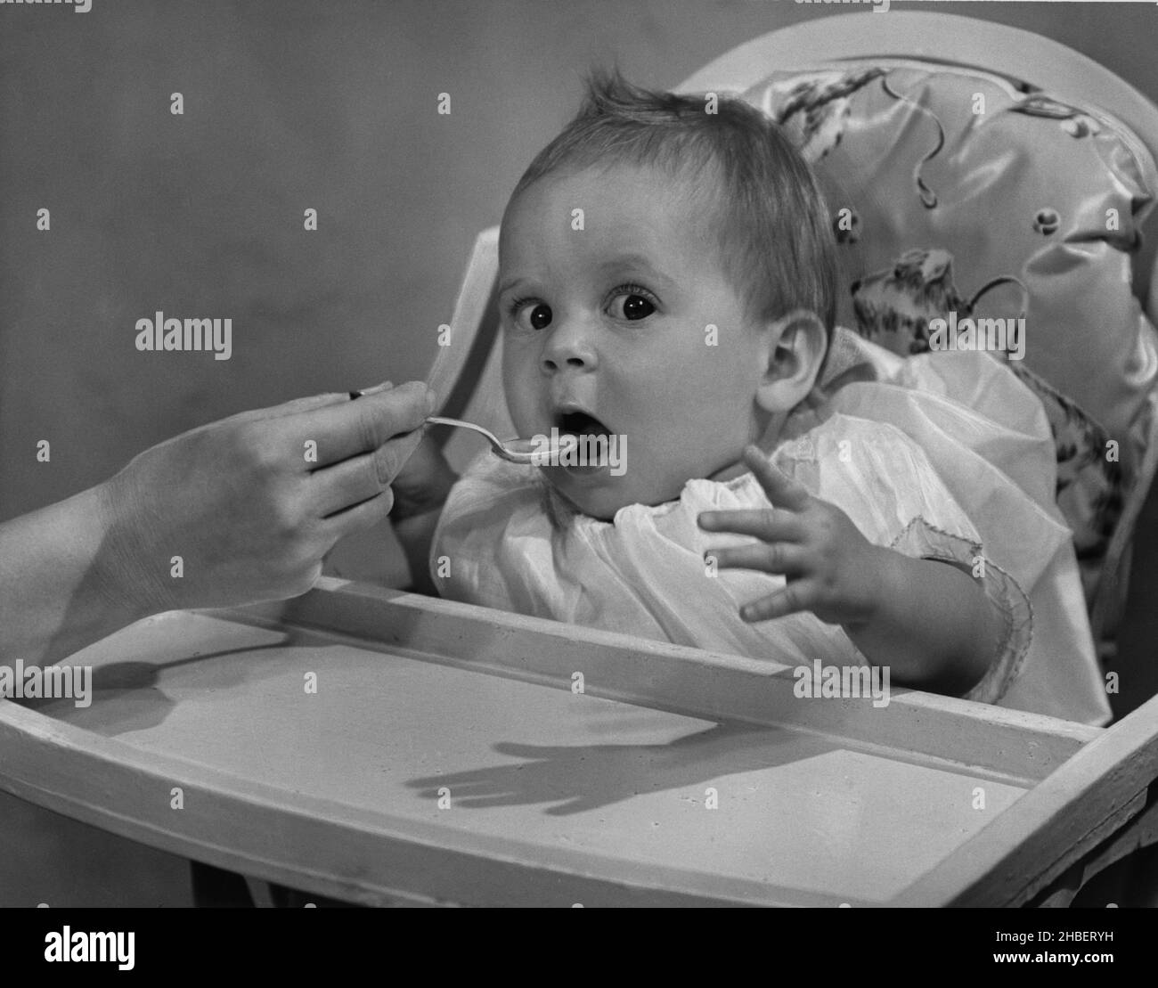 Bambina seduta in un hi-chair che viene alimentata a cucchiaio con uno sguardo sorpreso Foto Stock