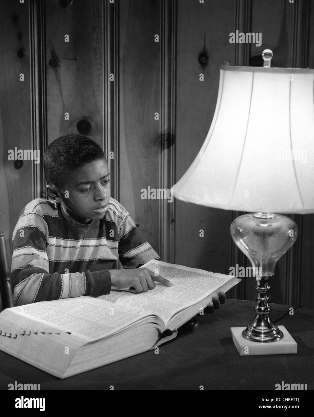 Ragazzo afroamericano seduto di fronte a una grande lampada con grande libro aperto e che punta a pagina Foto Stock