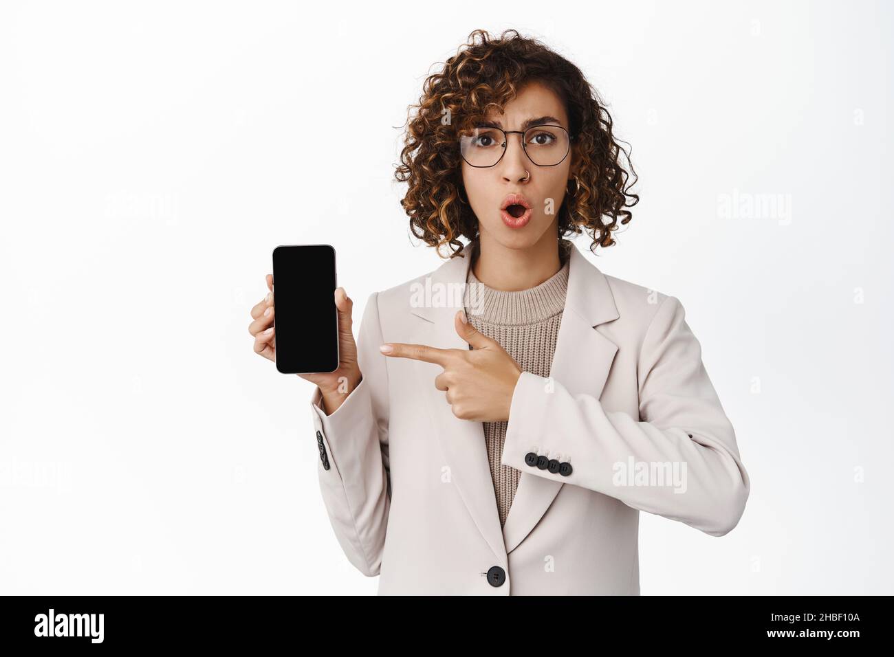 Scioccato imprenditore femminile in occhiali e vestito, puntando allo schermo del telefono cellulare con interessato, deluso volto espressione, sfondo bianco Foto Stock