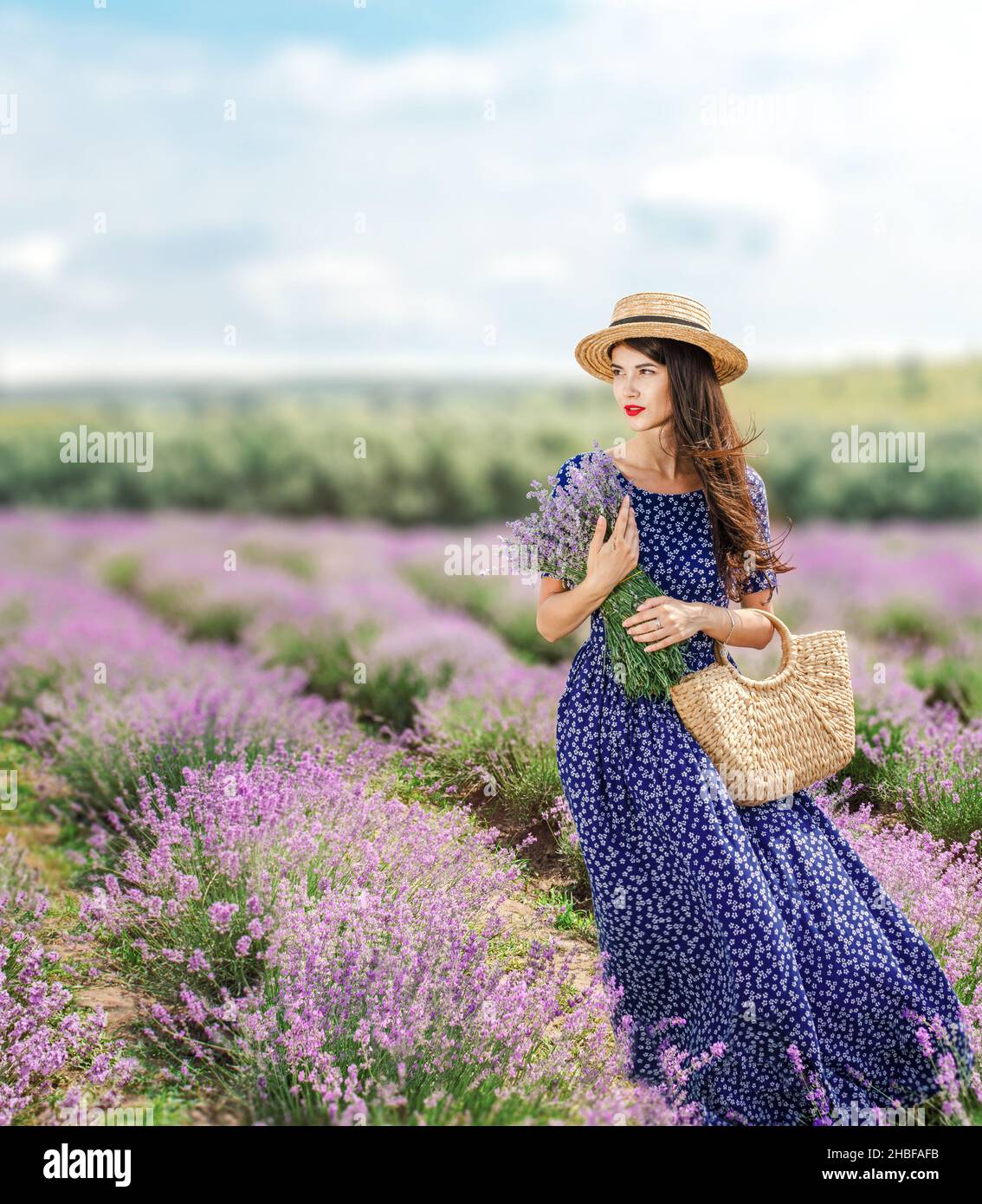 Donna bruna dai capelli lunghi in un campo di lavanda, in un lungo abito blu e cappello all'aperto in una giornata estiva. Foto Stock