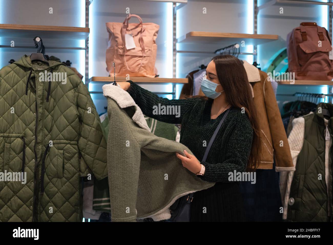 Cliente femminile in maschera che esamina cappotto elegante mentre sceglie i vestiti caldi nel negozio contemporaneo Foto Stock