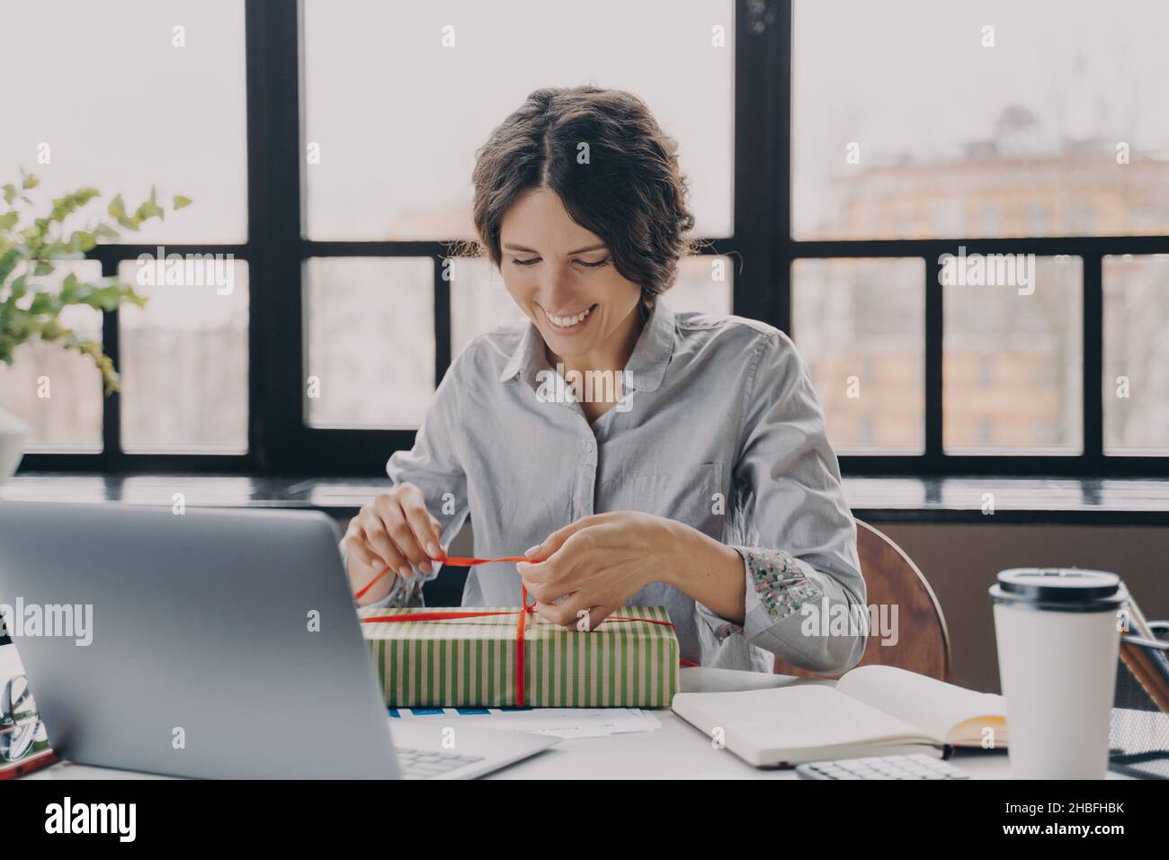 Giovane donna europea felice dipendente seduto sul posto di lavoro in ufficio e aprire la confezione regalo Foto Stock